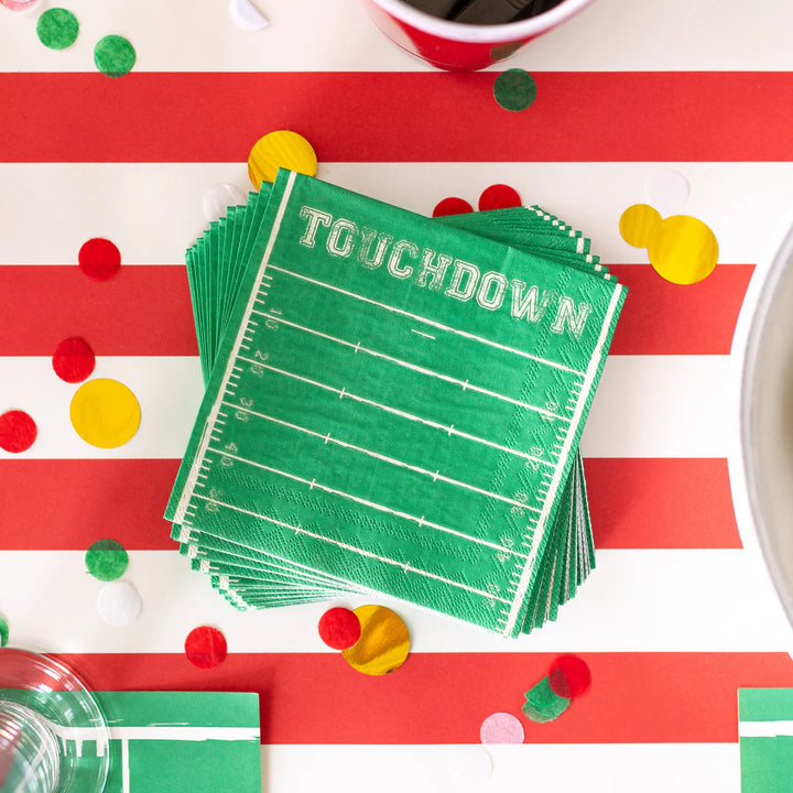 Stack of green 'Touchdown' paper plates on a red and white striped tablecloth with confetti.